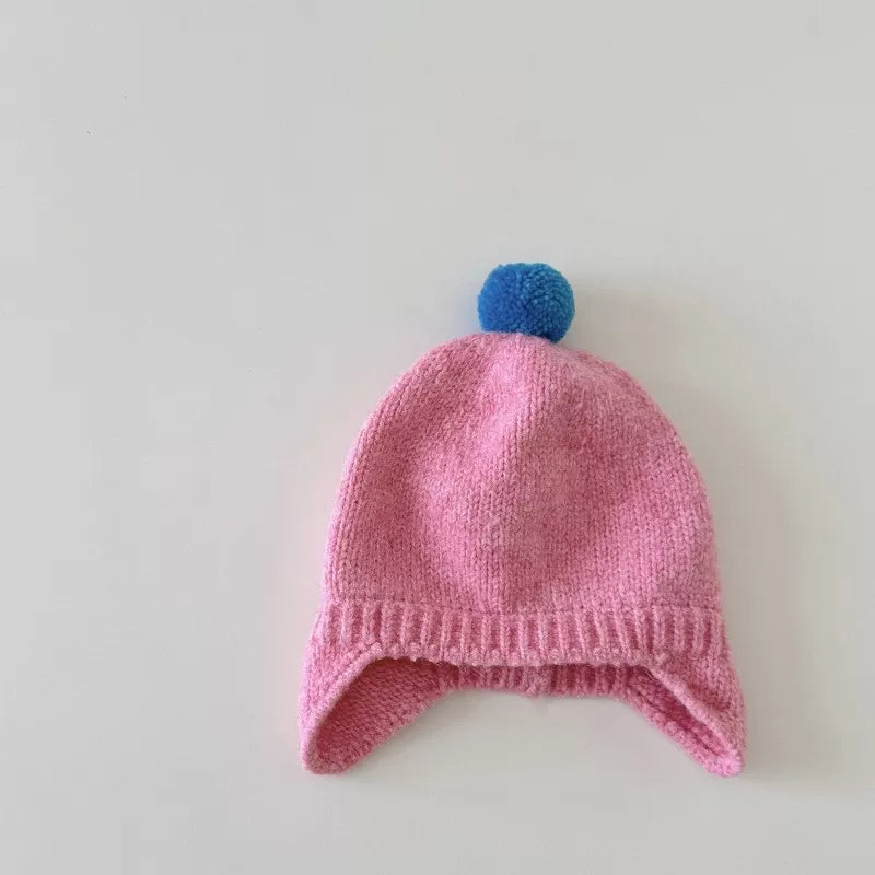 Rose pink knit baby earflap hat with blue pom-pom, flat lay