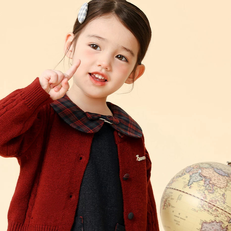 Little girl in a burgundy cardigan and navy plaid-collar pinafore dress waving