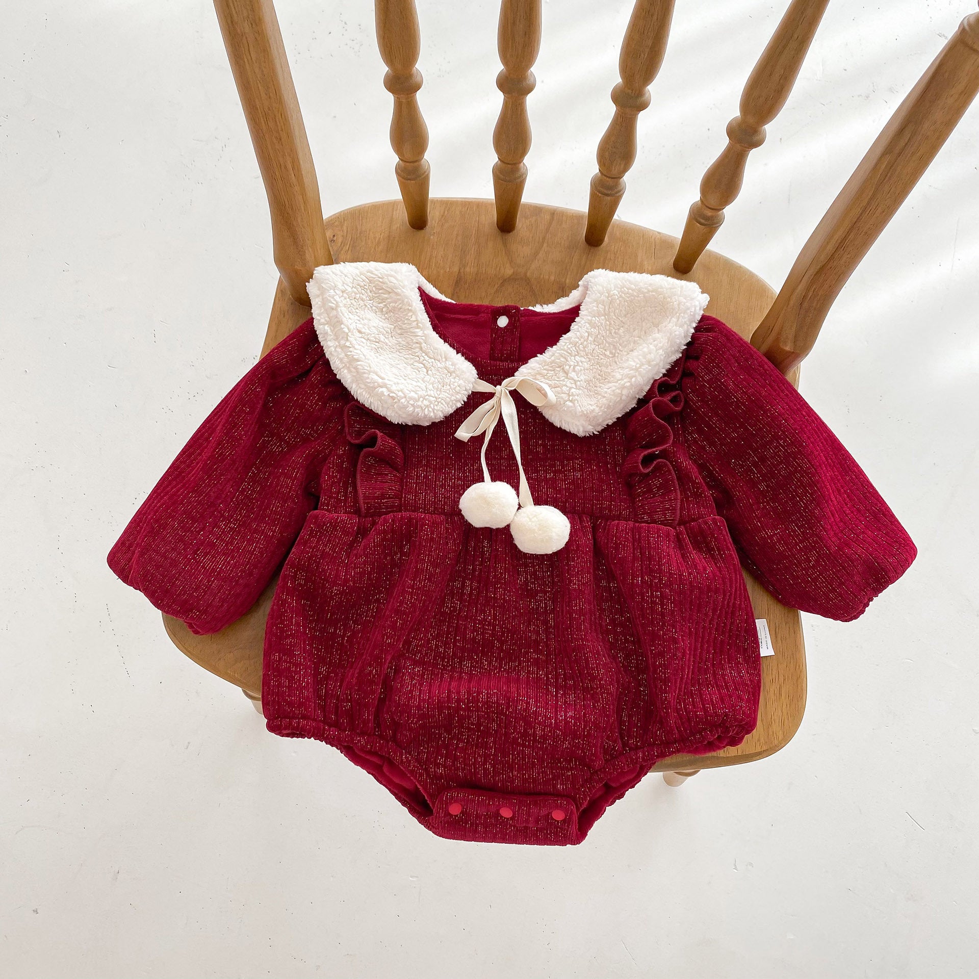 A luxurious red velvet baby romper with long sleeves, ruffled details, and a white faux fur Peter Pan collar adorned with a ribbon tie and two pom-poms. The romper is elegantly displayed on a wooden chair, highlighting its soft texture and festive design