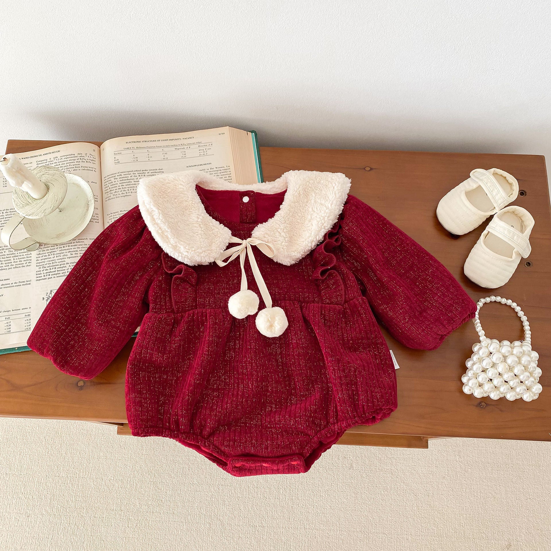 A festive red velvet baby romper featuring long sleeves, ruffled accents, and a soft white faux fur Peter Pan collar with a ribbon tie and pom-poms. Styled with matching accessories, including cream baby shoes, a pearl handbag, and displayed on a wooden table alongside an open book and a ceramic candle holder for an elegant and cozy setting.