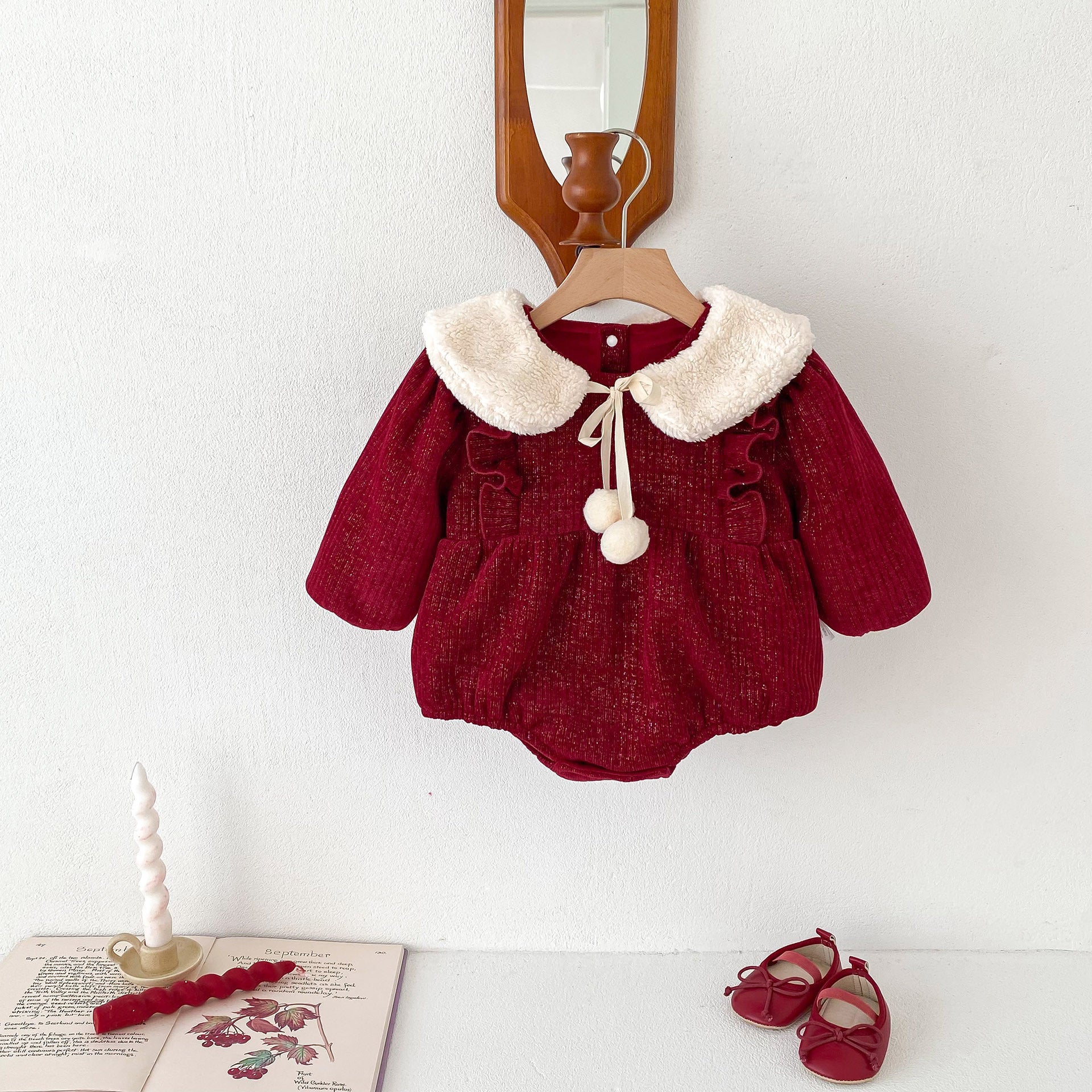 A red velvet baby romper with long sleeves, ruffled accents, and a plush white faux fur collar adorned with a ribbon tie and pom-poms. The outfit is displayed on a wooden hanger under a vintage-style mirror, styled with matching red baby shoes and a decorative setting that includes an open book, a twisted candle, and a red wax ornament for a festive and classic look.