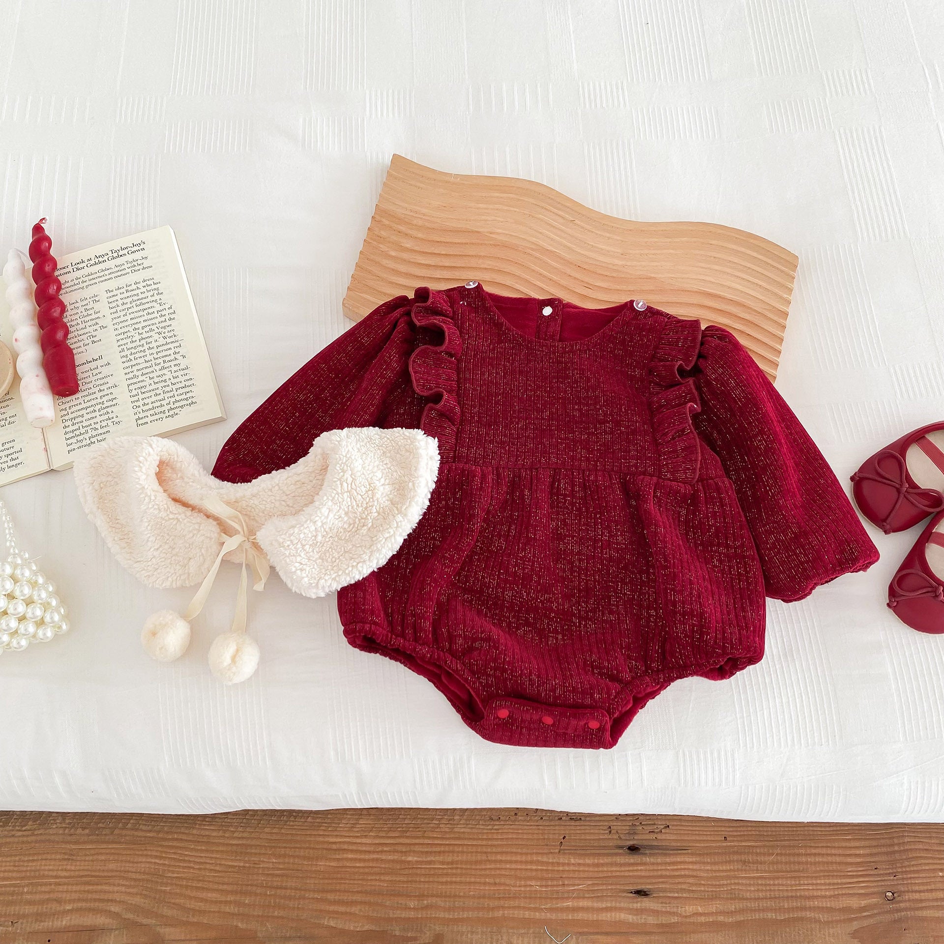 A luxurious red velvet baby romper with long sleeves and a plush white faux fur collar tied with a ribbon and pom-poms. The outfit is styled flat on a wooden surface with accessories, including an elegant pearl mini handbag, ivory baby shoes, and an open book for a timeless and sophisticated presentation