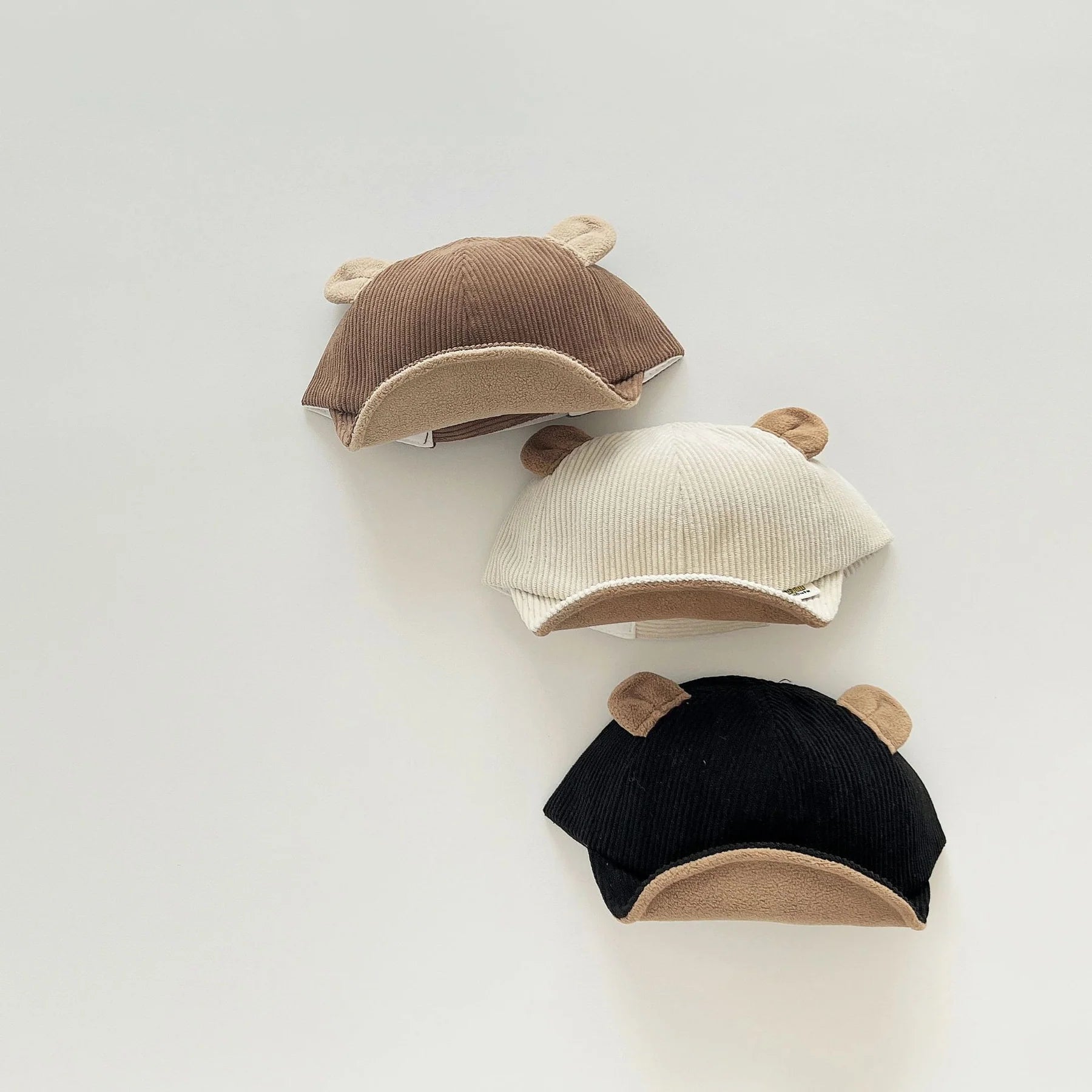 Set of three adorable children's caps in brown, cream, and black, each featuring bear ears on top. The caps have a soft ribbed texture and a curved, suede-like brim, adding a cute and cozy touch to any outfit. Displayed against a plain background, these caps are ideal for a stylish and playful accessory collection in a children's fashion store.