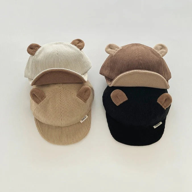 Four children's caps in a stack, featuring a ribbed texture with bear ear accents on top. The caps are in shades of beige, cream, brown, and black, each with a soft, curved brim. Ideal for adding a fun, playful touch to children's outfits, these caps offer a cozy and whimsical style. Displayed against a plain background, perfect for a children's fashion product showcase.