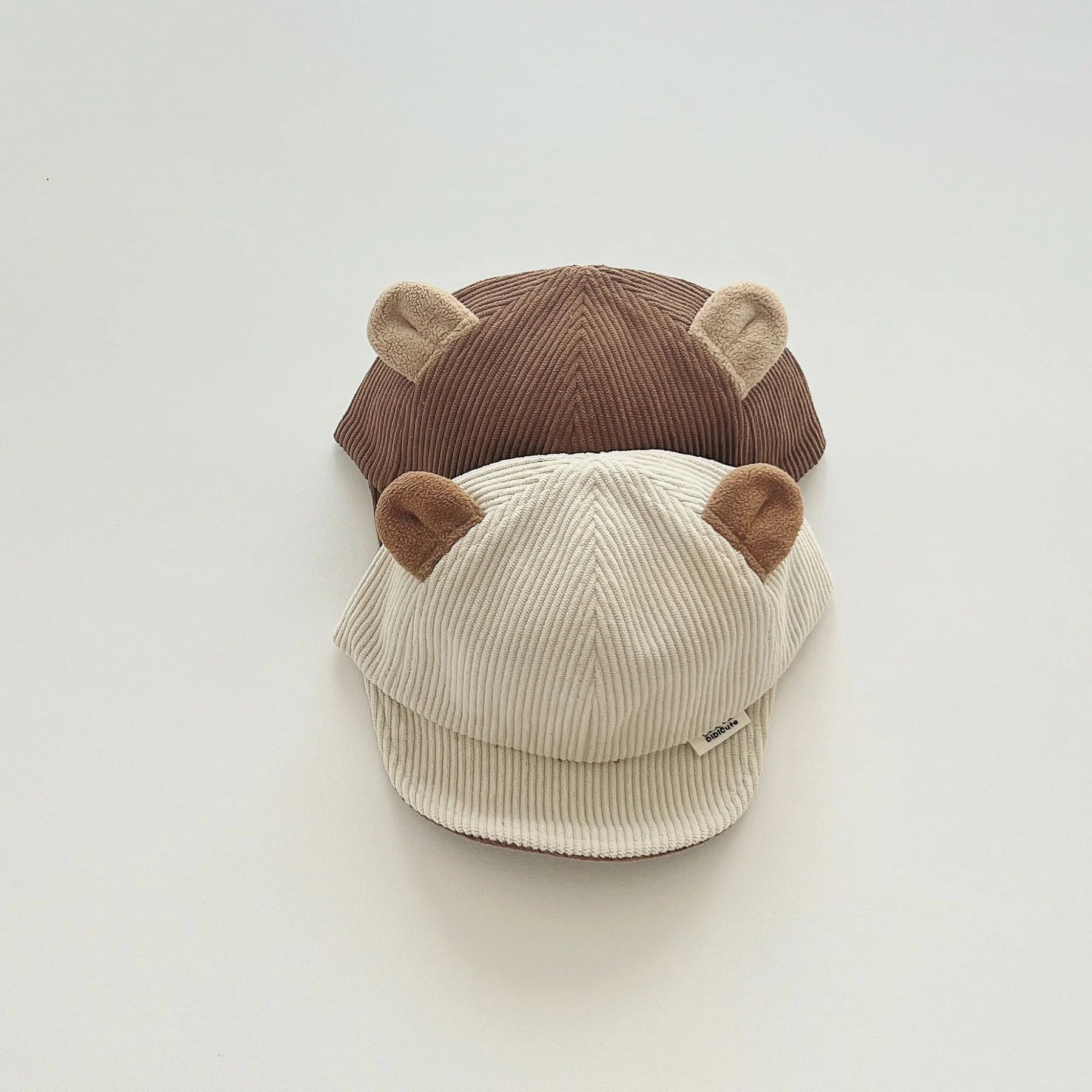 Two children’s caps with a ribbed texture, stacked together, each featuring soft, bear-like ears on top. The top cap is in a rich brown color, while the cap beneath is a light cream shade with brown ears. Displayed on a plain background, these caps combine playful design with cozy, textured fabric