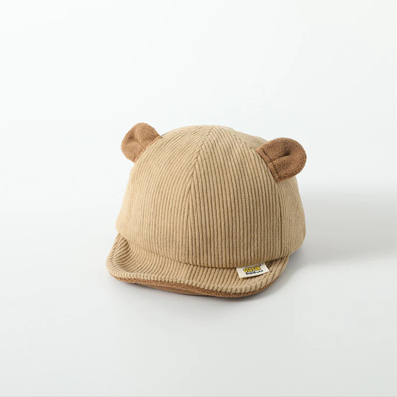 Children's beige cap with a ribbed texture and cute, bear-inspired ears on top. The cap has a small label on the front brim, adding a playful and cozy touch suitable for young children.