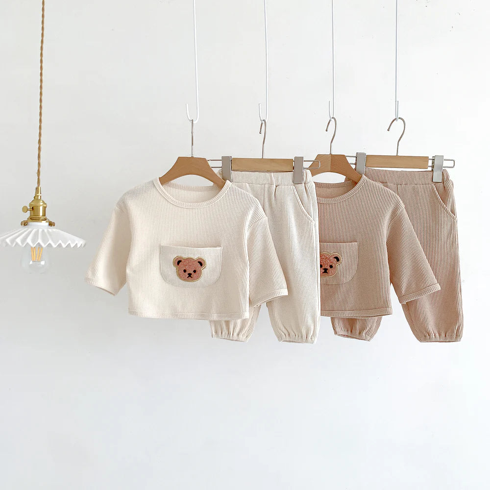 Adorable baby and toddler clothing set in neutral waffle-knit fabric, featuring two outfits with short-sleeve tops and matching pants, each with an embroidered teddy bear face on the front pocket. Displayed on wooden hangers against a minimalist background, with a charming vintage-style hanging lamp adding warmth to the scene. The outfits come in beige and light brown tones, perfect for cozy casual wear in spring and fall.