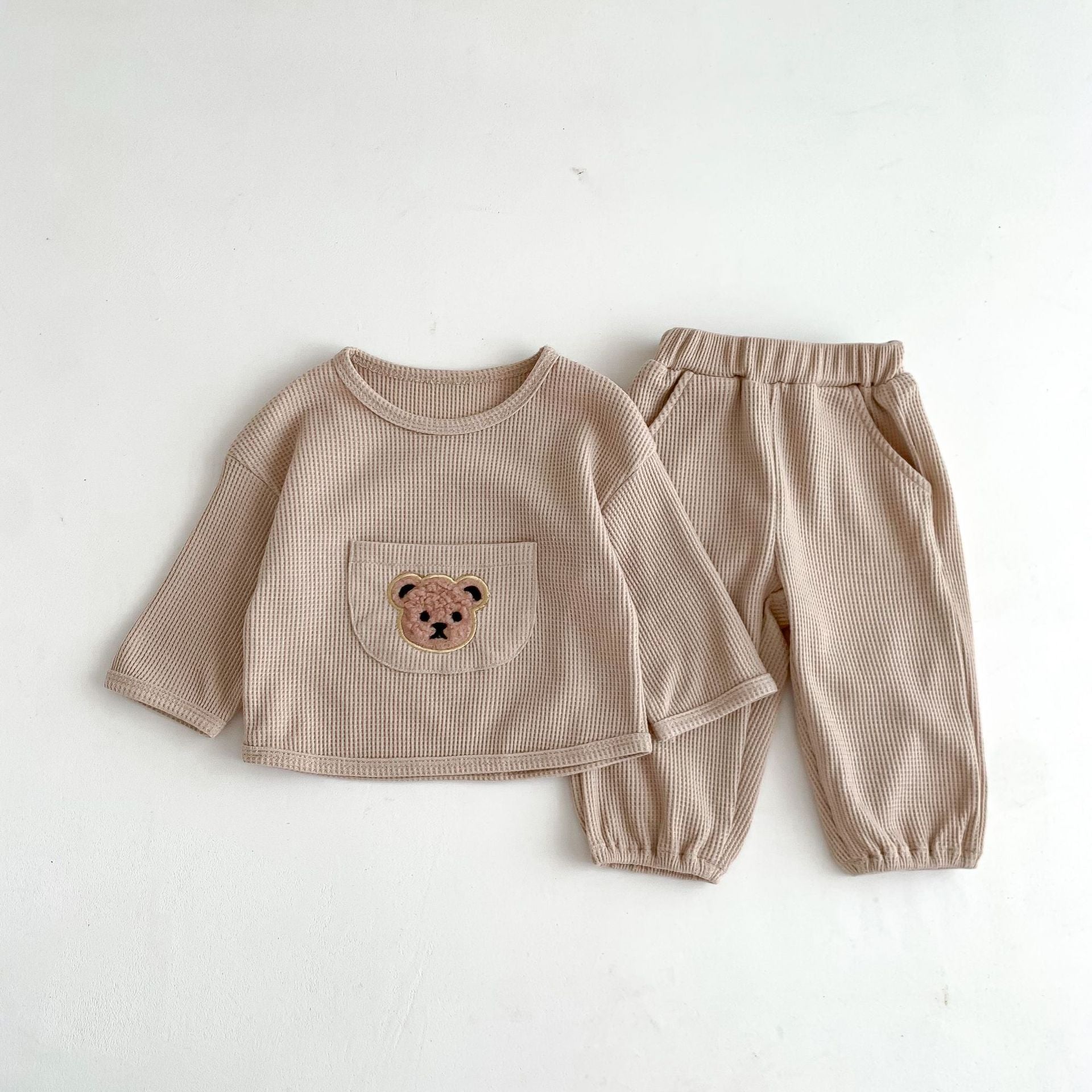 Cute baby and toddler outfit set in a soft beige waffle-knit fabric, featuring a long-sleeve top with an embroidered teddy bear face on the front pocket, paired with matching pants. The pants have an elastic waistband and cuffed ankles for a comfortable fit, making this set perfect for cozy everyday wear in cooler weather.