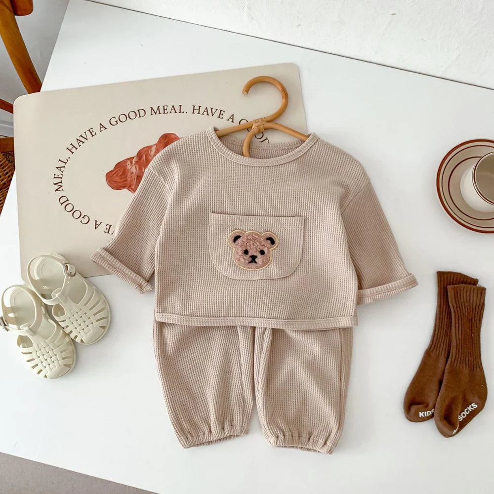 Alt text: "Adorable baby outfit set in beige waffle-knit fabric, featuring a long-sleeve top with a front pocket decorated with an embroidered teddy bear face, paired with matching pants. The set is displayed with complementary accessories, including white jelly sandals, brown ribbed socks, and a minimalistic ceramic cup, creating a cozy, stylish look. A decorative placemat with the phrase 'Have a good meal' adds a playful touch to the setup