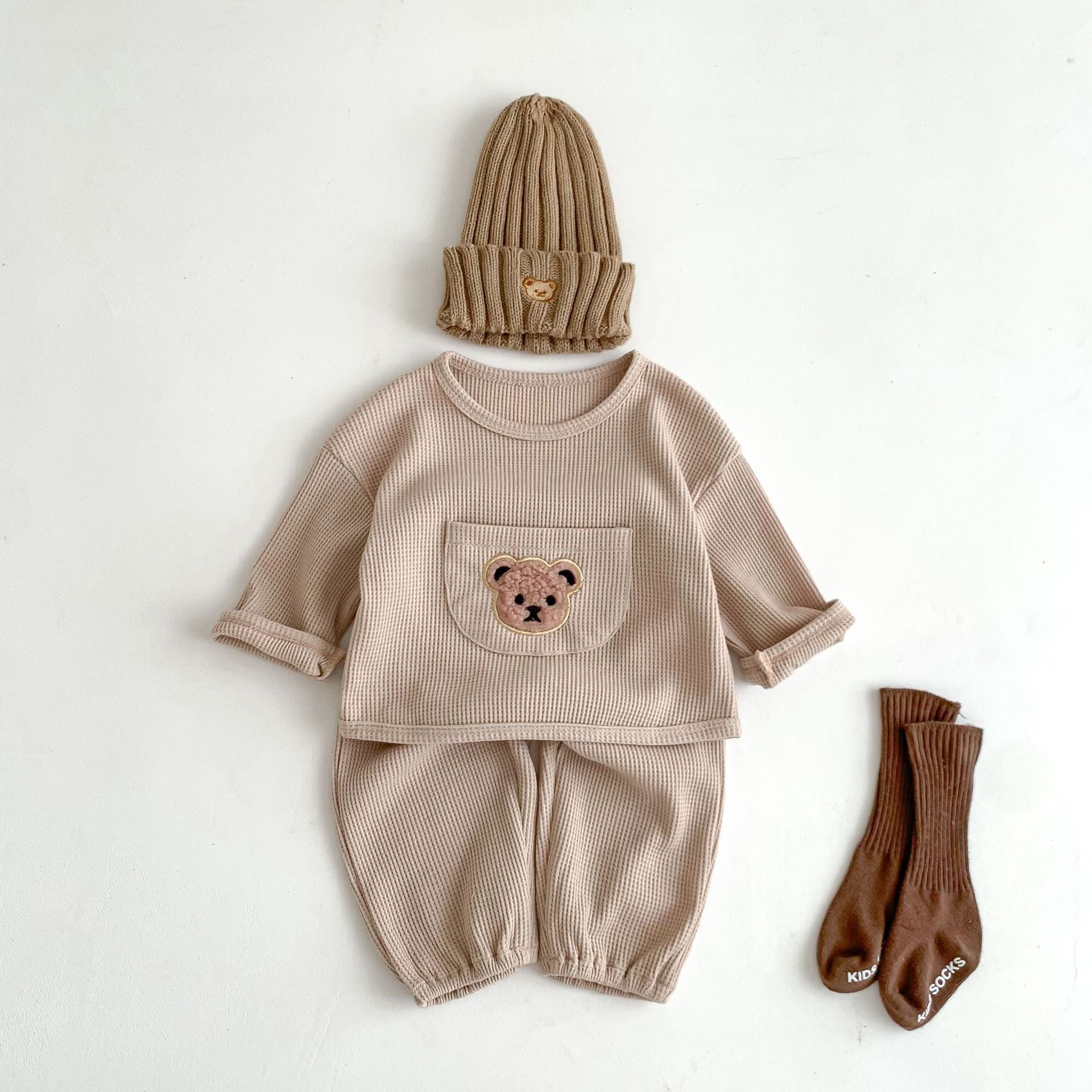 "Cozy baby outfit set in beige waffle-knit fabric, including a long-sleeve top with a front pocket embroidered with a teddy bear face and matching pants with elasticated cuffs. Styled with a coordinating ribbed beanie with a bear patch and a pair of brown ribbed socks, this outfit is perfect for a comfortable, warm look in cooler weather. The soft, neutral tones and adorable bear design make this set an ideal choice for gender-neutral baby loungewear.