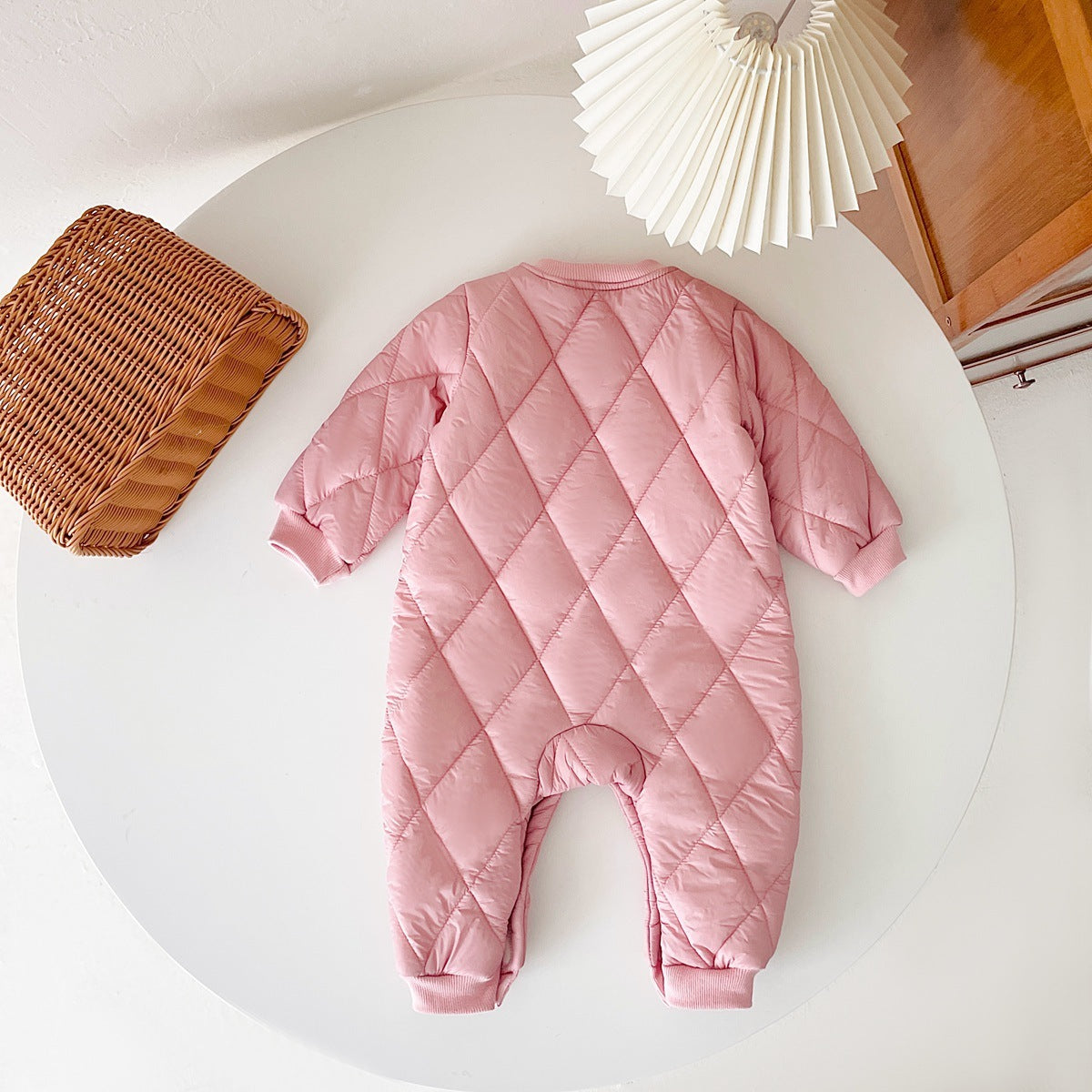 Back view of a pink, quilted baby jumpsuit displayed on a round white table. The jumpsuit features a diamond quilt pattern, long sleeves, and ribbed cuffs for added comfort and warmth. A wicker basket and a white pleated lampshade are placed nearby, adding a cozy aesthetic.