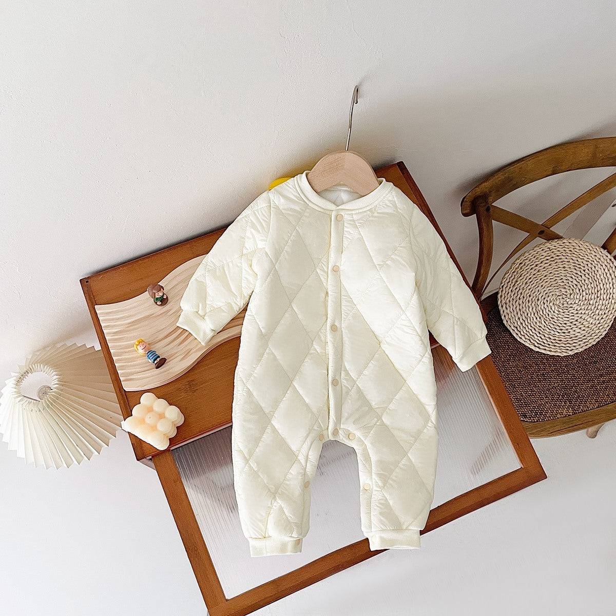 Cream-colored quilted baby jumpsuit with fleece lining, laid on a wooden display table. The jumpsuit features diamond-pattern stitching, snap buttons along the front, and long sleeves, designed for warmth and comfort. The cozy, padded design is ideal for cold weather. The background includes a decorative lamp and small toys, adding a warm, homely feel