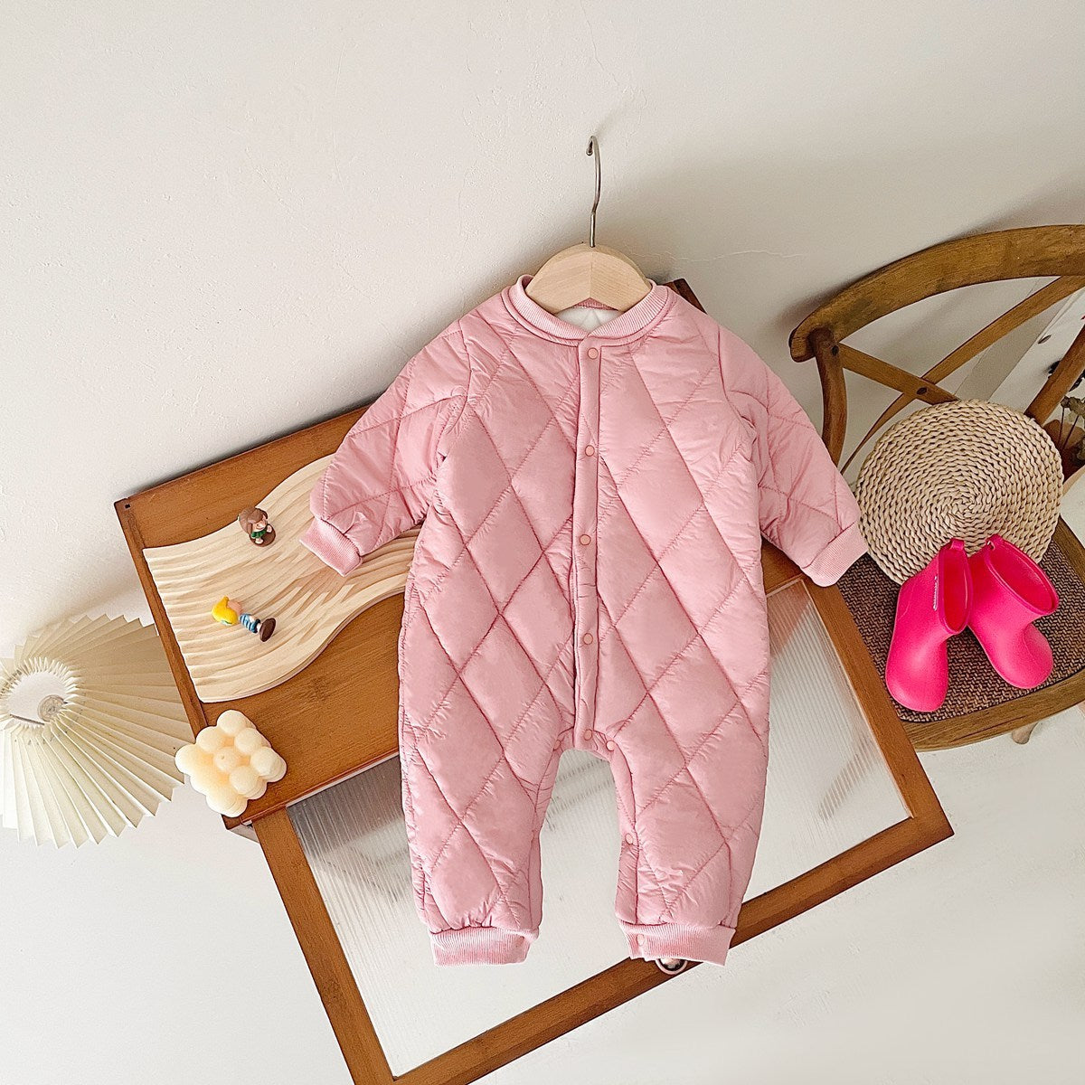 Pink quilted baby jumpsuit with fleece lining, hanging on a wooden display table. The jumpsuit features a diamond-patterned design, snap buttons down the front, and long sleeves, providing warmth and comfort for cold weather. Nearby are decorative elements, including a modern lamp, toys, and a pair of small red rain boots, creating a cozy and playful setting.