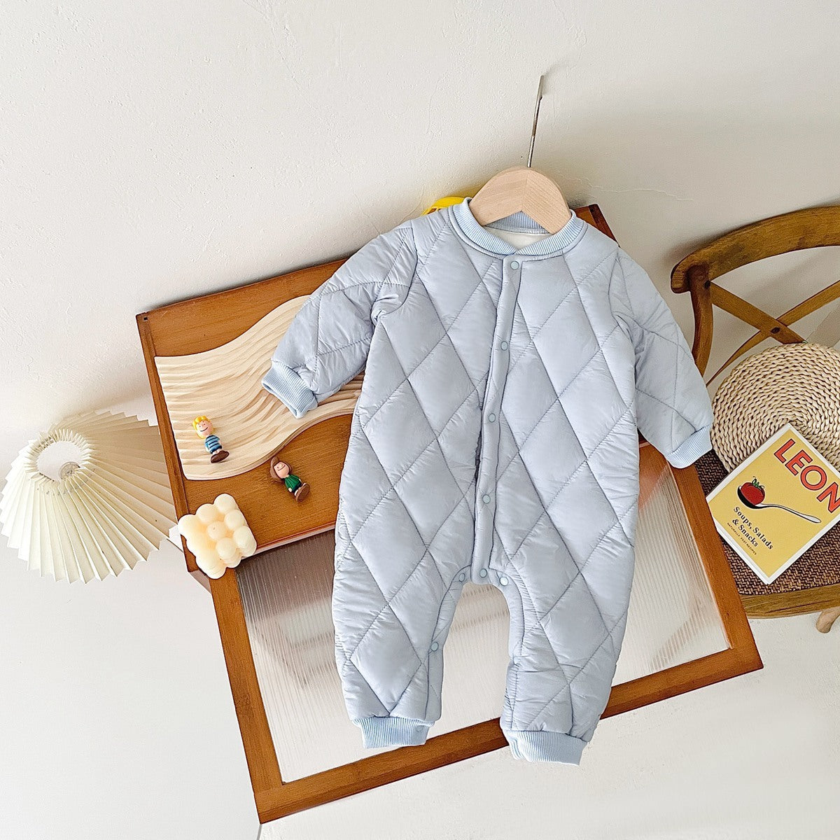 Light blue quilted baby jumpsuit with fleece lining, displayed on a wooden tabletop. The jumpsuit has a cozy diamond-patterned design, long sleeves, and snap buttons down the front, ideal for keeping babies warm in colder weather. Surrounding decor includes a small book, wooden toys, and a decorative lamp, creating a warm and inviting scene.