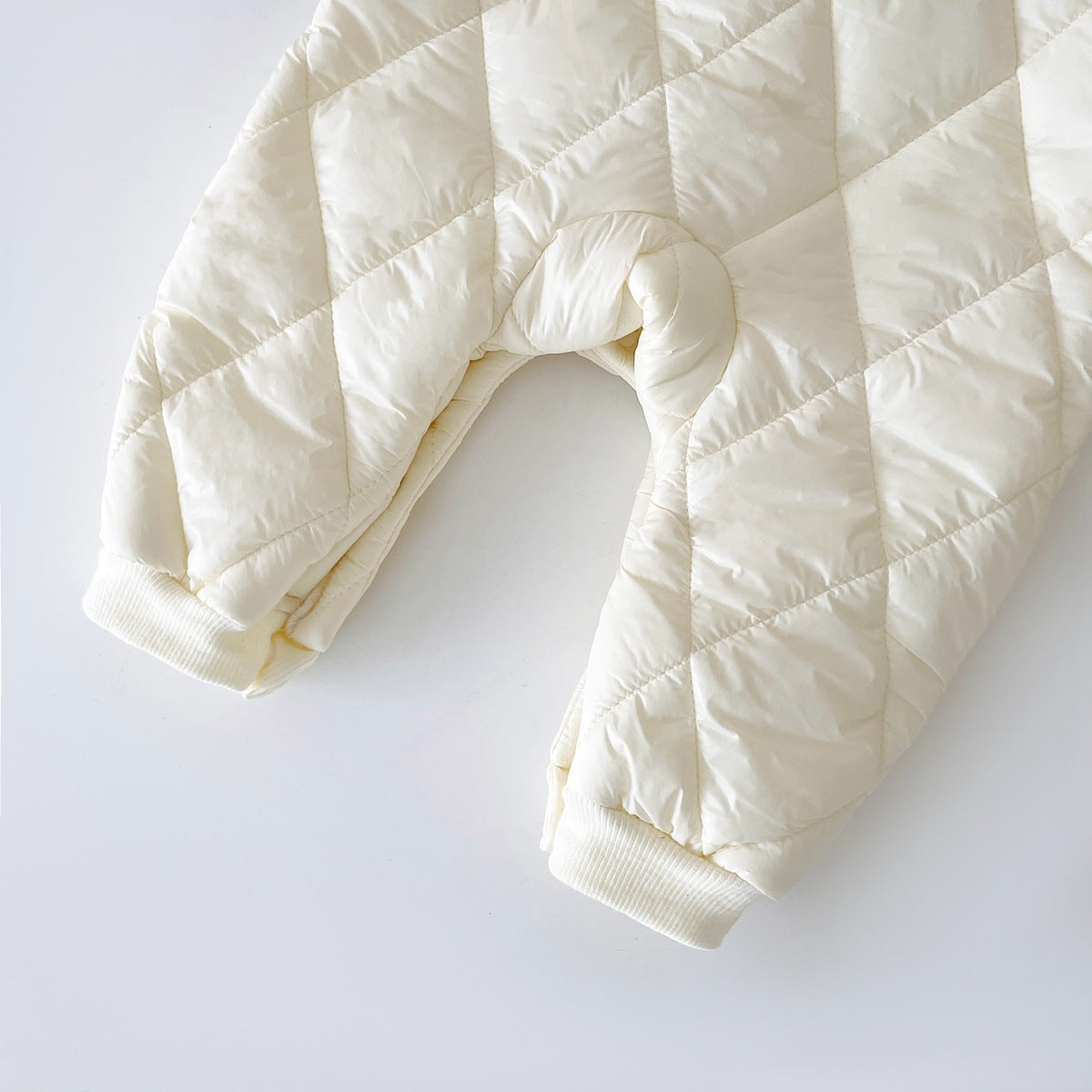 Close-up of the lower part of a cream-colored, quilted baby jumpsuit with a soft fleece lining. The jumpsuit features a diamond pattern and elastic cuffs at the ankles, providing a snug and warm fit for colder days.