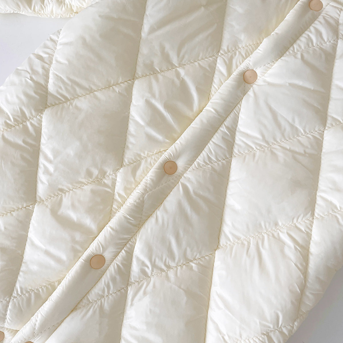 Close-up of a cream-colored, quilted baby jumpsuit with a diamond pattern and snap buttons along the front. The jumpsuit has a soft, padded texture, ensuring warmth and comfort for the baby during colder seasons.