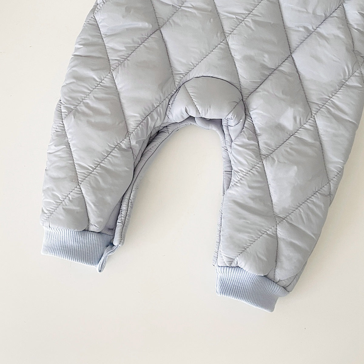 Close-up of a light gray, quilted baby jumpsuit with a diamond pattern, showing ribbed cuffs at the ankle for added comfort and warmth. The jumpsuit is padded, offering insulation for cold weather, and features soft inner lining visible at the ankle openings.