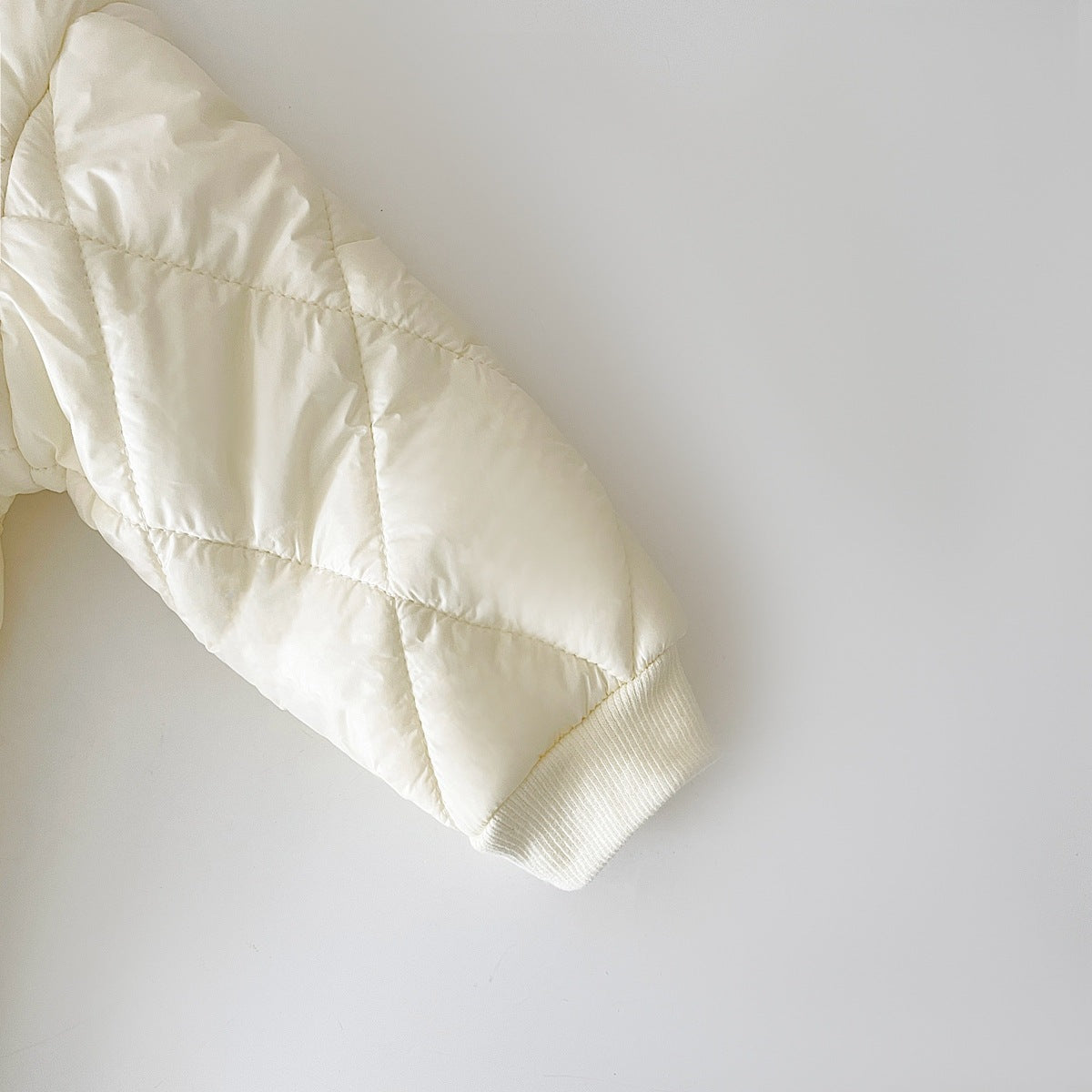 Close-up of the sleeve of a cream-colored, quilted baby jumpsuit with a diamond pattern, showing a ribbed cuff for a snug fit at the wrist. The quilted padding offers warmth, making it suitable for cold weather.