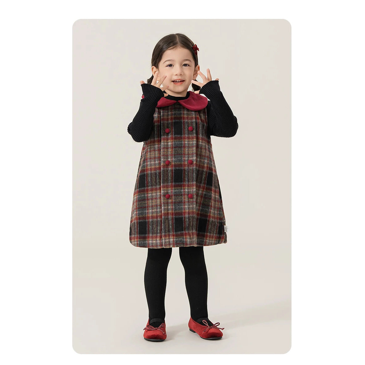 Toddler girl smiling in a plaid pinafore dress with black sweater and red shoes