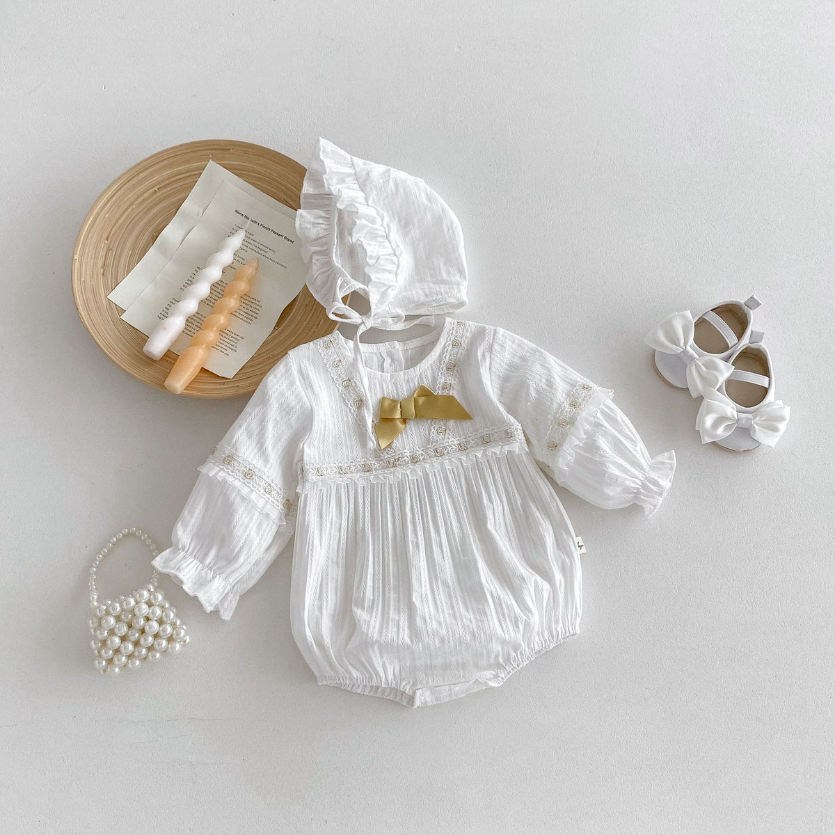 White muslin baby romper with lace trim, gold bow and ruffle bonnet, flat lay