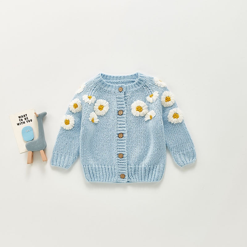 Charming baby cardigan in a soft pastel blue, embellished with embroidered white daisy flowers with yellow centers on the shoulders and sleeves. The button-up front features wooden buttons, adding a rustic and classic feel to this cozy knit. Accompanied by a cute blue giraffe toy and a small card with a friendly message, this setup showcases the cardigan's playful and stylish details,