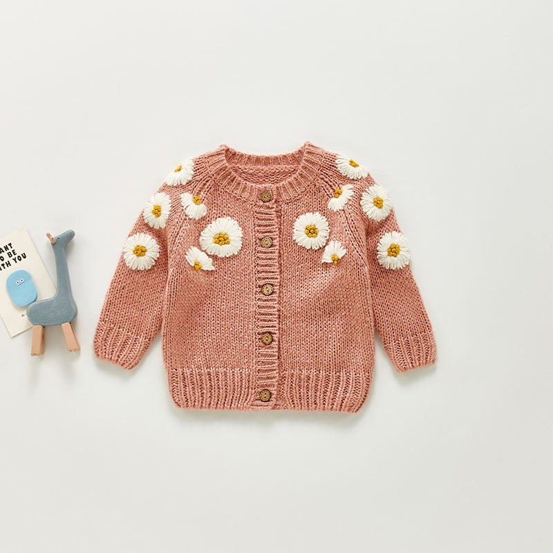 Adorable baby cardigan in a warm pink hue, adorned with embroidered white daisies with yellow centers across the shoulders and sleeves, creating a cheerful and cozy design. The cardigan features a button-up front with wooden buttons, adding a classic, rustic touch. Paired with a small blue giraffe toy and a cute card, this setup highlights the cardigan's charming and playful floral details,